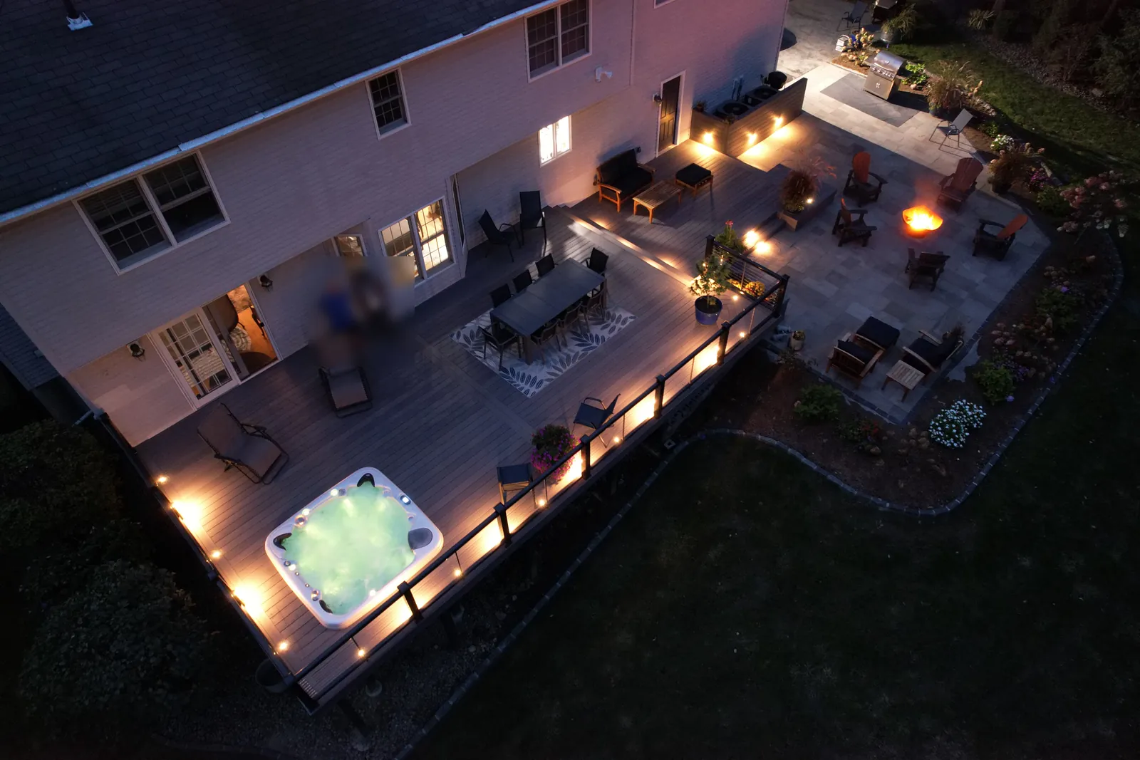 Composite deck with spa, dining and lounge areas beside a lit paver patio and fire pit at night