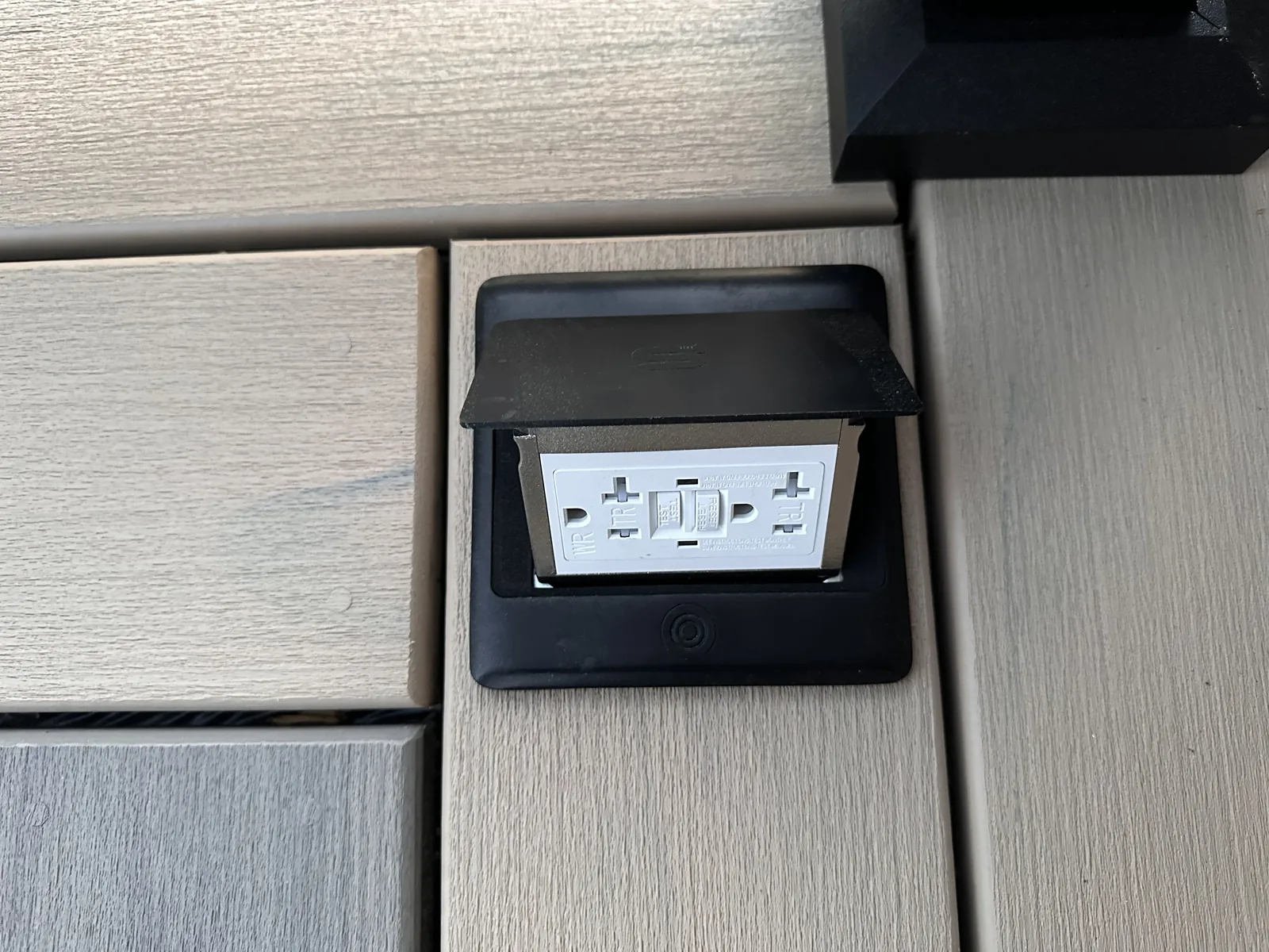 Weather-rated pop-up GFCI outlet set flush into a composite deck surface, lid open — the in-deck power detail demonstrated at the Messco display
