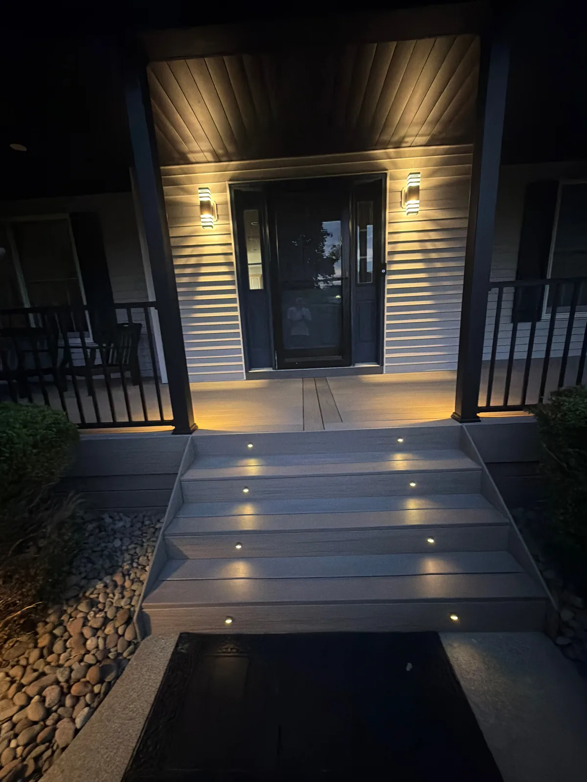 Front porch at night — riser lights in the steps, sconces lit, black aluminum railings