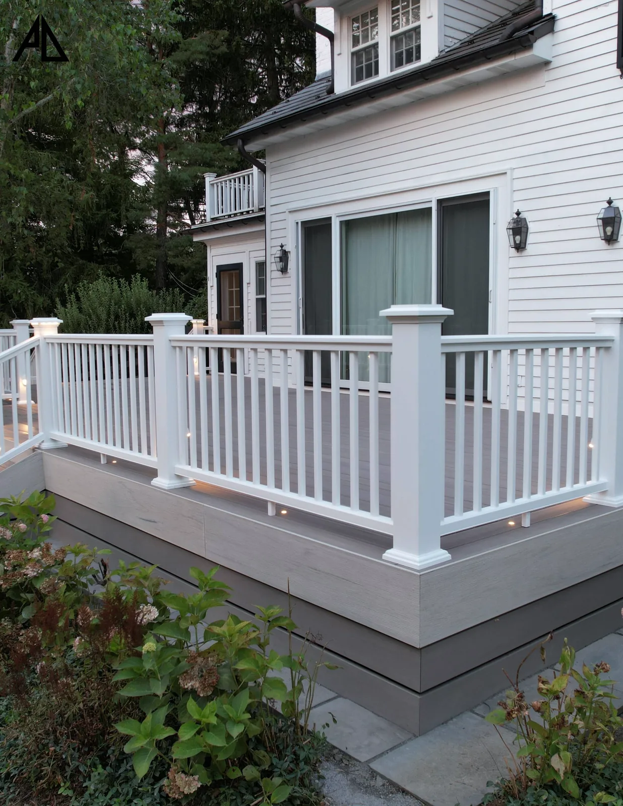 TimberTech deck corner at dusk with white square-baluster railing, lit fascia, and hydrangeas at grade