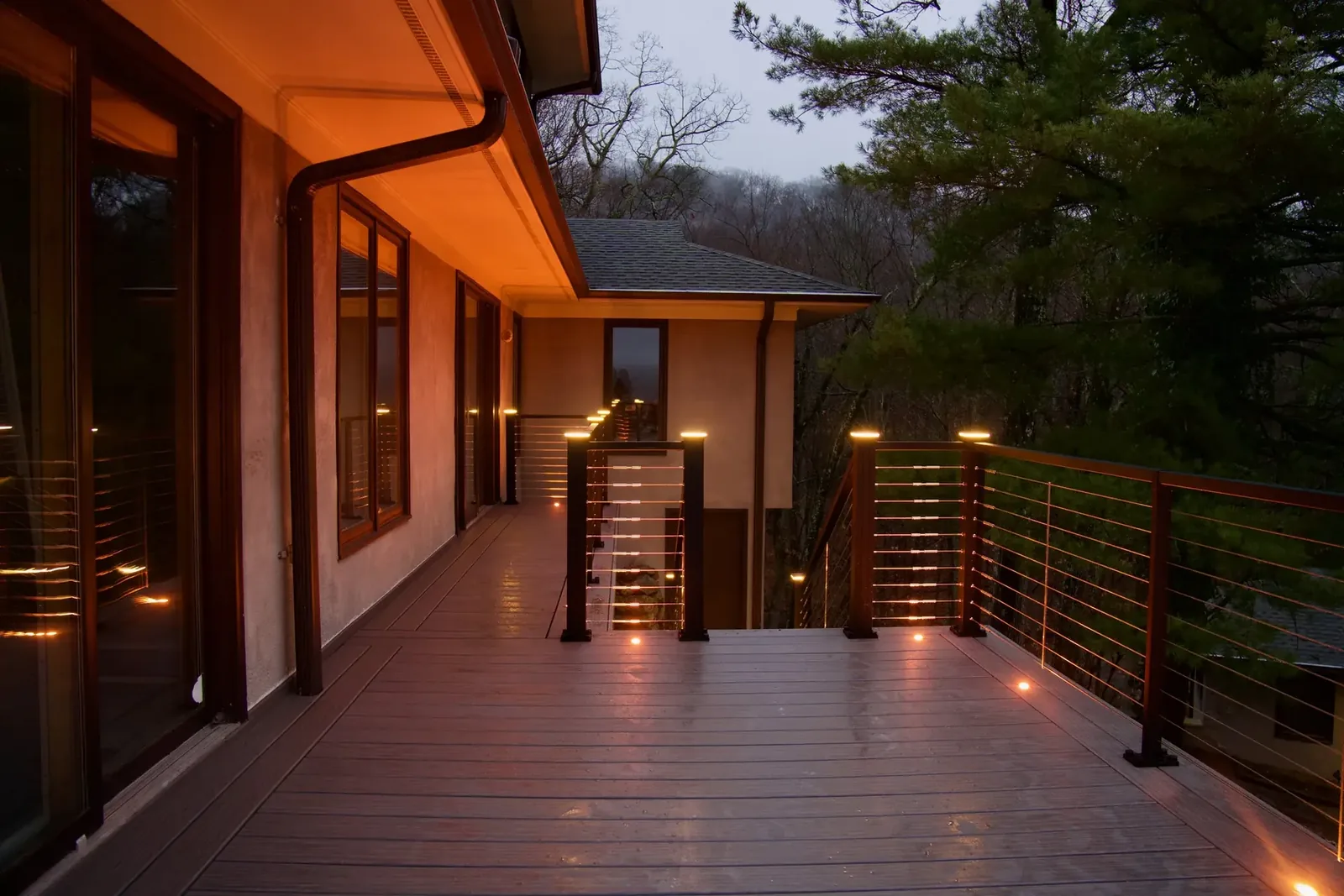 Composite deck at dusk with signature under-rail LED glow, illuminated cable railing, post-cap lights, and flush deck lighting over misty Hudson Valley woods — Pinnacle Decking Haven Lighting