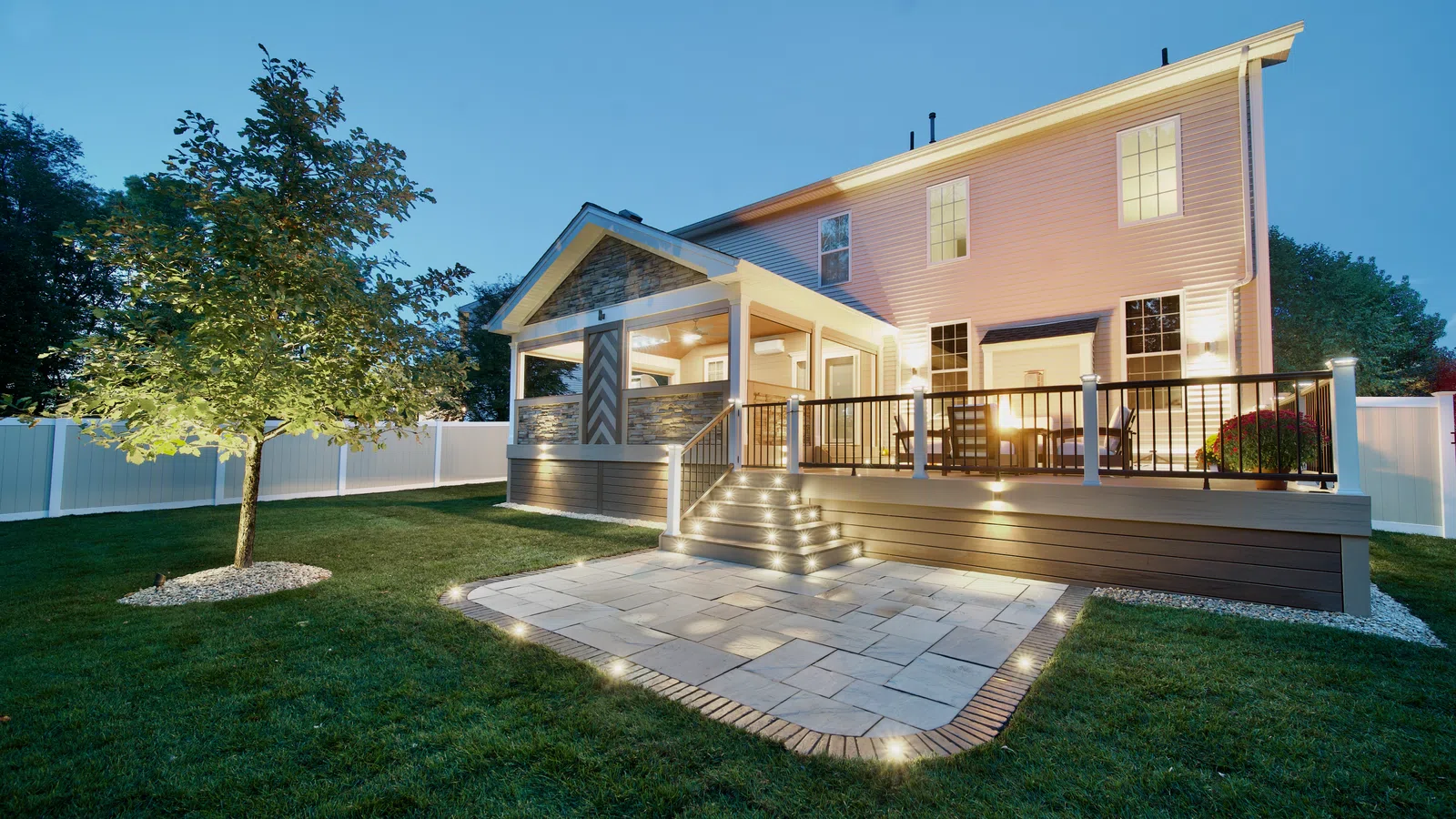 Twilight backyard with lit deck, covered porch, and paver patio