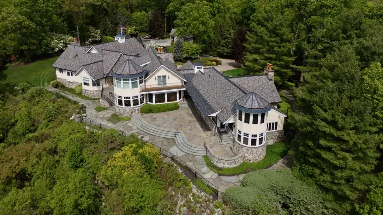 Aerial of a cream hilltop house with a stone patio, curved stone steps, and wooded slopes