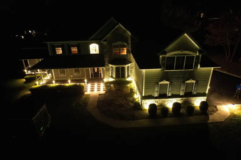 Aerial night view of stone-and-siding house with landscape lighting along porch steps, foundation and front walkway