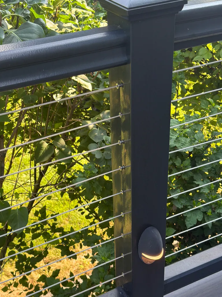 Close-up of cable railing post with integrated light fixture, sunlit foliage behind