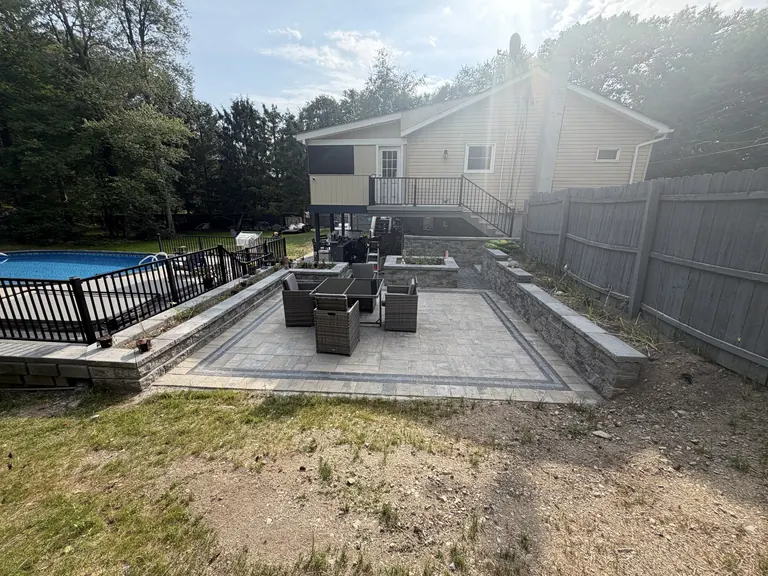 Wide view of raised paver patio with seating area, stone retaining walls and elevated deck with stairs behind the house