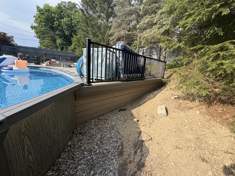 Composite deck skirting and black aluminum railing along above-ground pool edge, wooded backyard