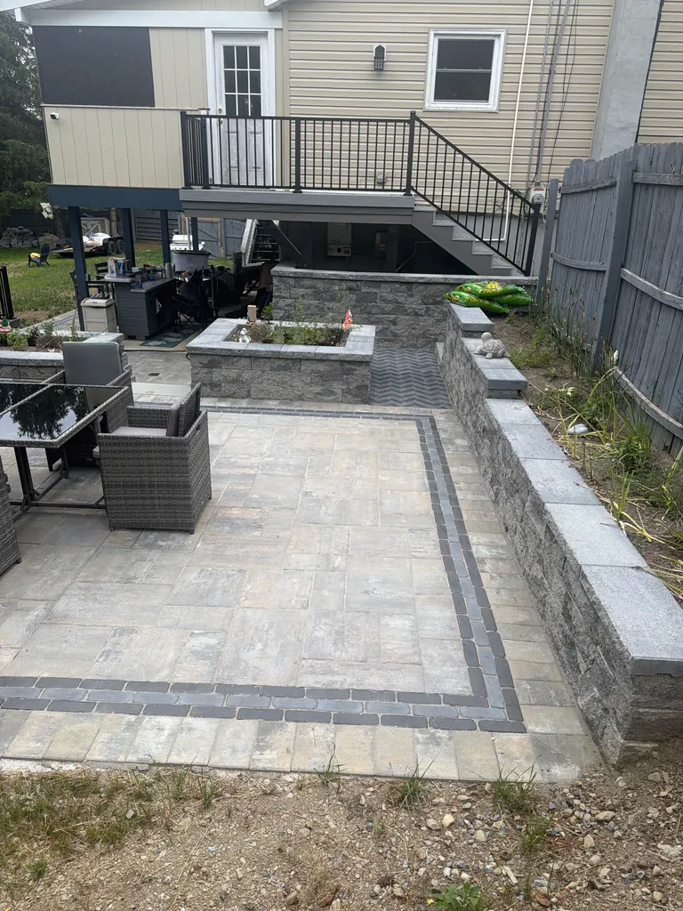 Finished paver patio with double-border inlay, retaining wall planter, herringbone accent, and black-rail deck stairs above