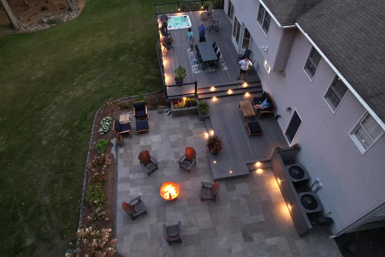 Twilight aerial of full deck, hot tub, dining, and stone patio with fire pit and string lighting