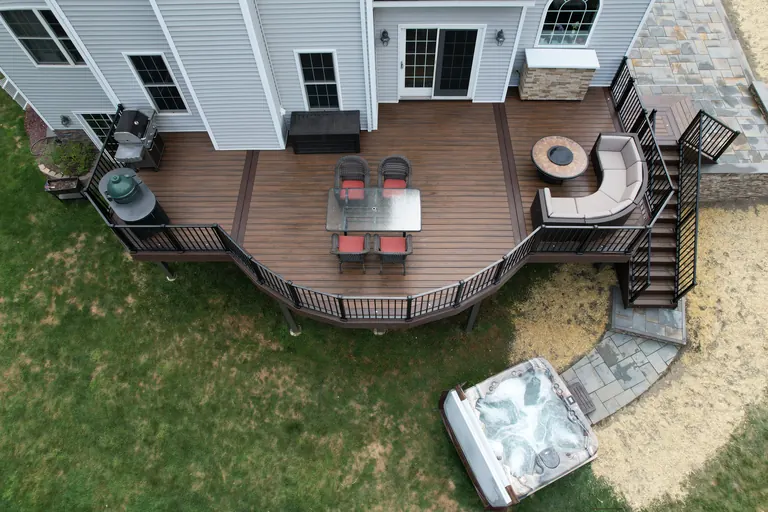 Curved composite deck with black cable railing, built-in bench seating, fire table, and a hot tub below on a stone patio, Dutchess County, NY