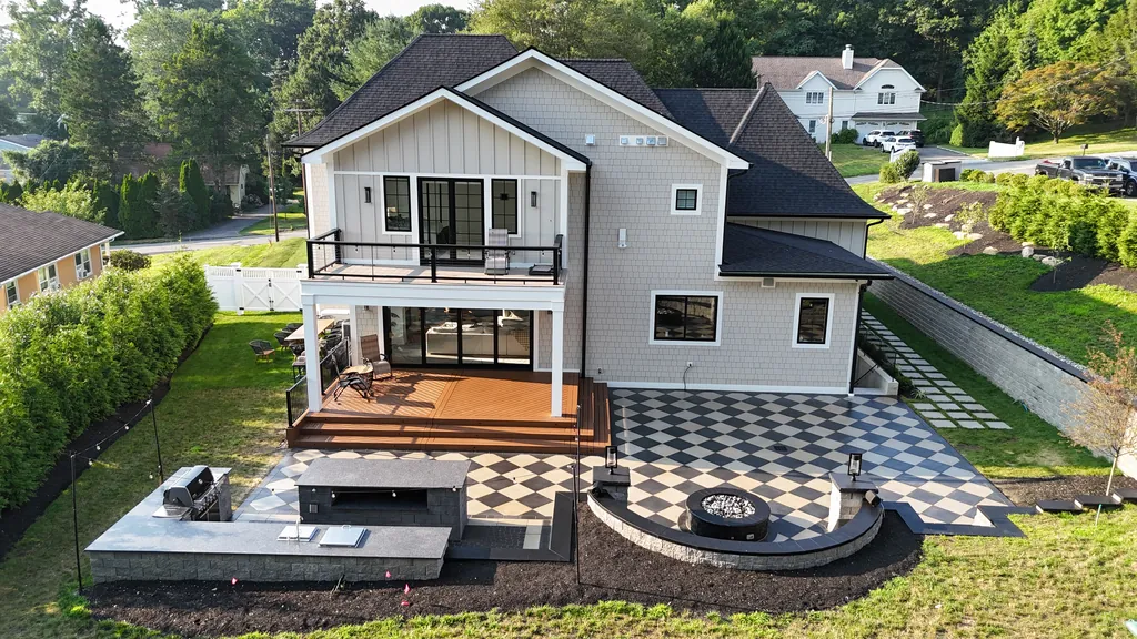 Aerial view of a deck above a black and cream checkerboard paver patio with a built-in grill and a round fire pit