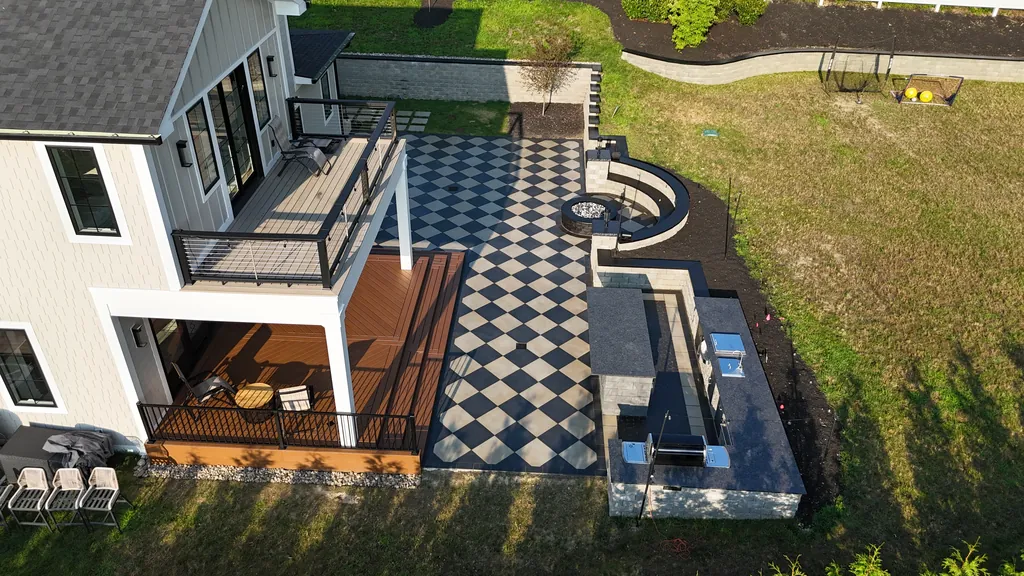 Aerial view of a two-level deck with black railing over a checkerboard paver patio, outdoor kitchen and fire-pit seat wall