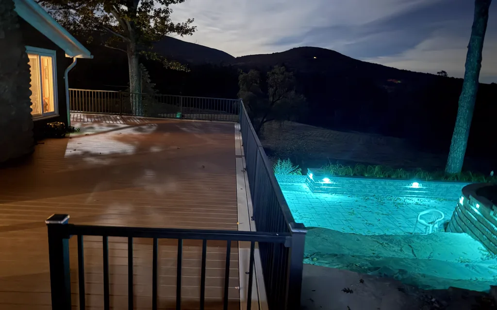 Deck with black railing beside a lower patio lit by blue-green wall lights, mountains at dusk