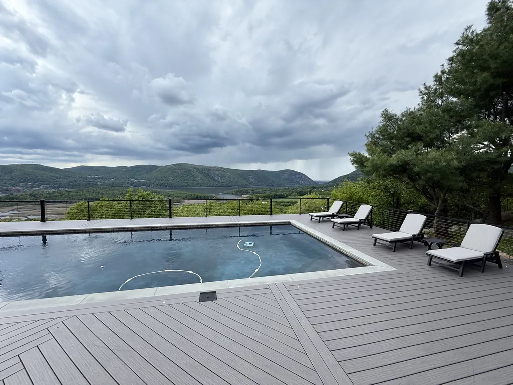 Gray deck around a pool with lounge chairs, mountains and river beyond
