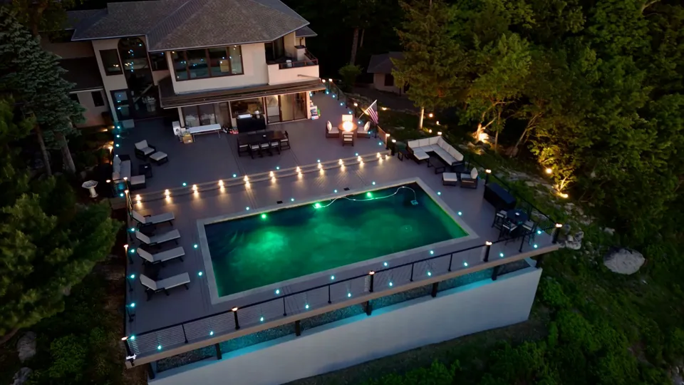Aerial view at dusk of a lit pool deck, deck lights and a fire table outside a hillside home
