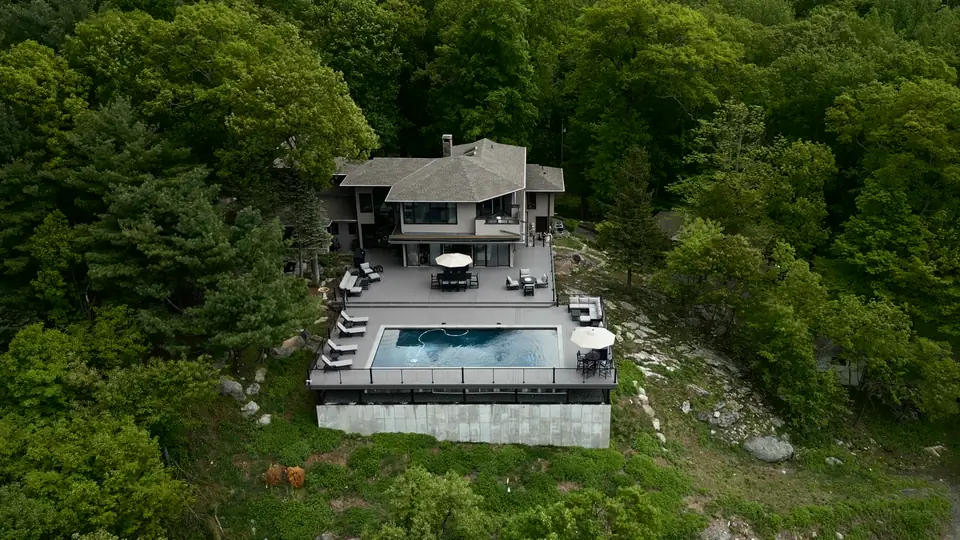 Aerial view of a hillside home with a pool deck, black cable railing and outdoor dining among green trees