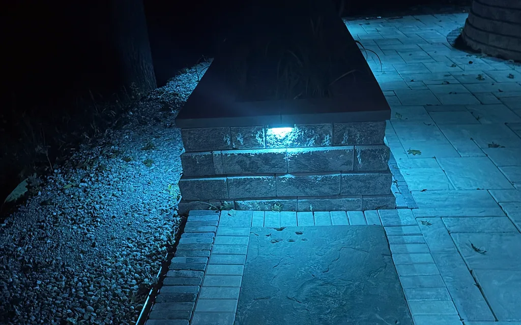 Wall light glowing in a stone planter wall beside a paver walkway at night