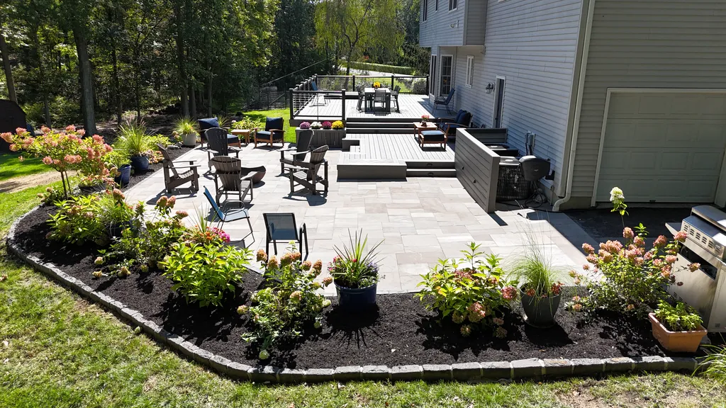 Stone patio with Adirondack chairs and garden beds, a deck beyond